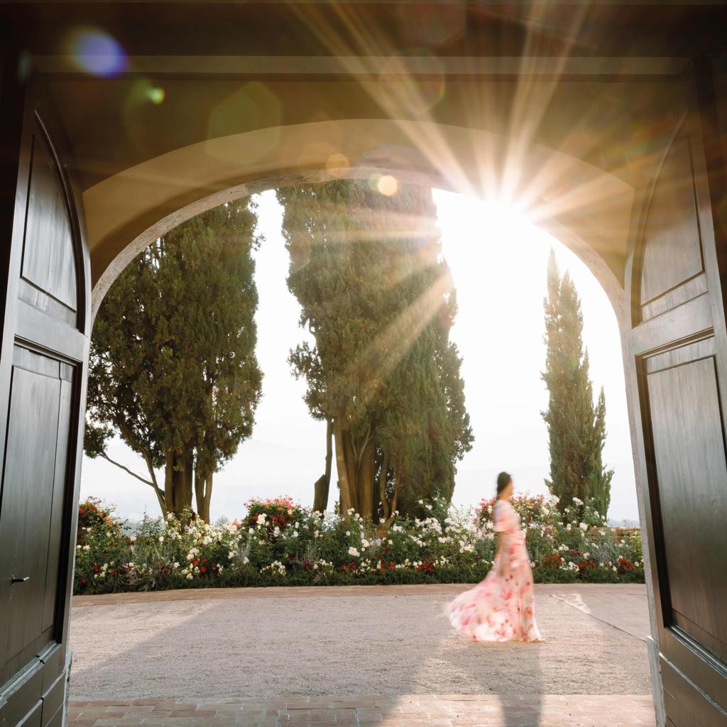 A woman in a pink dress walks through open arched doors into bright morning sun